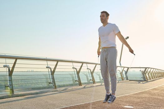 Full length portrait of a muscular build European middle aged man, fit athlete doing jumping exercises, cardio training with skipping rope on the urban city bridge early in the morning on a summer day