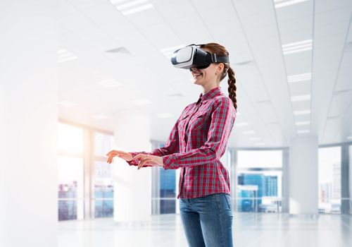 Young woman wearing virtual reality helmet in modern office interior. Mixed media