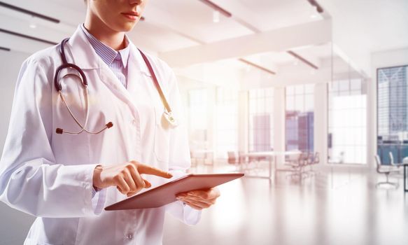 Cropped image of young woman doctor touching tablet screen with finger while standing inside bright medical office building with sunlight on background. Medical industry concept