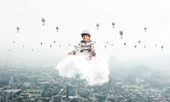Young little boy keeping eyes closed and looking concentrated while meditating on cloud in the air with cityscape view and flying aerostats on background.