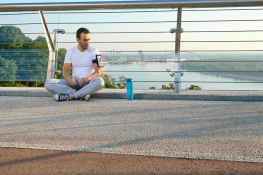 Handsome Caucasian man in sportswear sitting on a city bridge and checks a mobile application with monitoring heart rate and calories burned during a workout on his phone in a smartphone holder