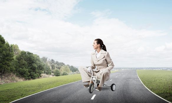 Beautiful young woman riding children's bicycle on asphalt road. Businesslady in white business suit cycling small bike outdoor. Nature landscape with green grass. Professional career start concept