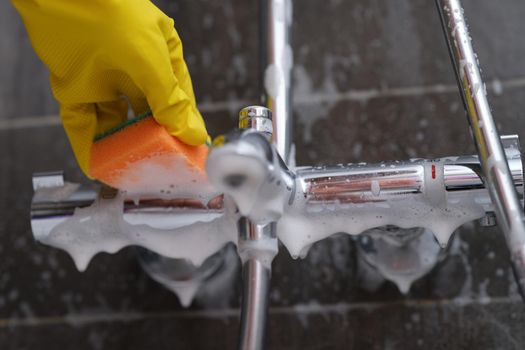 Cleaning lady in rubber glove washes faucet in bathroom with sponge with foam closeup. Cleaning company services concept