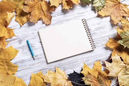 Spiral sketchbook and pen lies on vintage wooden desk with bright foliage. Flat lay with autumn leaves on white wooden surface. Blank notepad for drawing with copy space. Creative space and artwork.