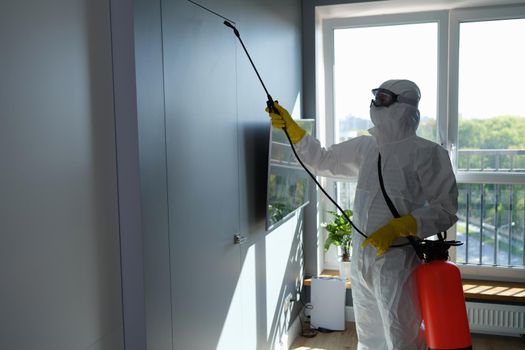 Disinfection to destroy viruses. Worker in protective suit and face mask sprays chemicals indoors concept