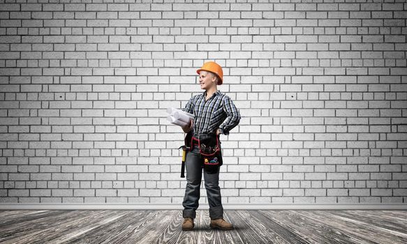 Attractive female architect in hardhat standing with technical blueprints. Portrait of young worker in checkered blue shirt and jeans standing in empty room. Interior design and renovation service.