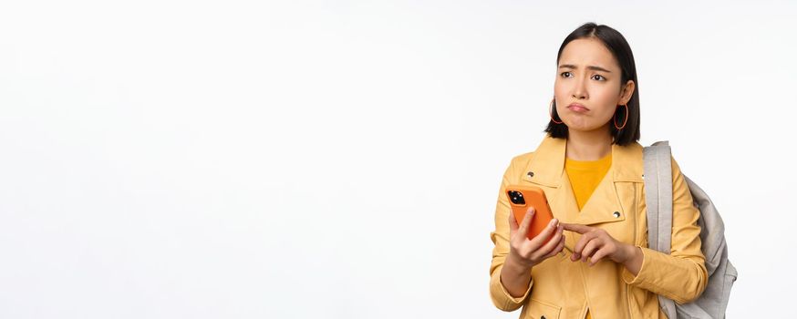 Image of happy girl traveller, tourist with backpack looking at smartphone, using mobile phone route app, standing over white background. Copy space