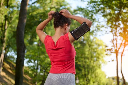 Rear view of a young determined sportswoman wearing a smartphone holder, tying ponytail, getting ready for run along the city park outdoor