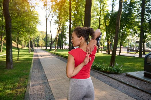 Muscular fit athlete, middle aged confident sportswoman raising arms up, stretching out and warming up her body before morning jog along the city park on a beautiful summer sunny day