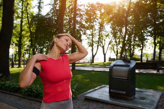 Hispanic sporty woman in tight red t-shirt with smartphone holder standing on the city park doing stretching exercise, enjoying morning workout or warming up her body before morning jog