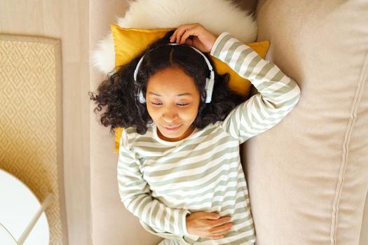 Happy and calm African-American female smiling while lying down on sofa in living room and listening to music in headphones. Relaxing and chilling at home. Showing positive emotion. Horizontal above