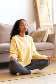 African-American woman in sportswear bare feet sitting in yoga lotus position on mat in living room while meditating and doing breathing therapy. Concept of mindfulness and stress relief