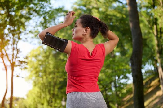 Rear view of a sportswoman wearing a smartphone holder, tying ponytail, getting ready for run along the city park outdoor