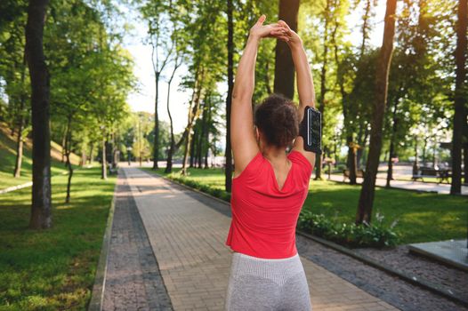 View from the back to a muscular fit athlete, sportswoman raising arms up, stretching out and warming up her body before morning jog along the city park on a beautiful summer sunny day