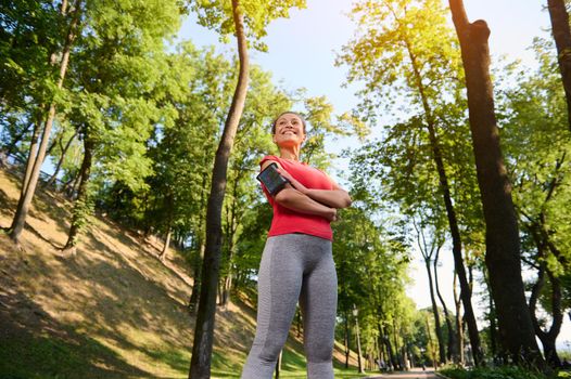 Bottom view of a confident 30 years old pretty woman in sportswear standing in a forest city park with her arms crossed and smiling with a beautiful toothy smile during her morning run or workout