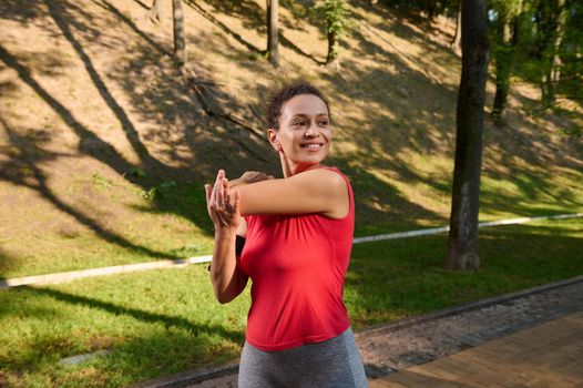 Determined charming middle aged Hispanic woman stretching out her arms while exercising outdoors on a warm summer day. Sport, fitness, body weight training, weight loss concept for healthy fit body