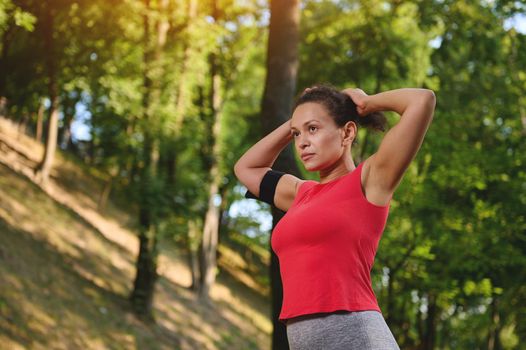 Beautiful African American woman, determined female athlete tying ponytail, preparing for jog along the city park on a sunny summer day. Sport, body weight training and slimming concept