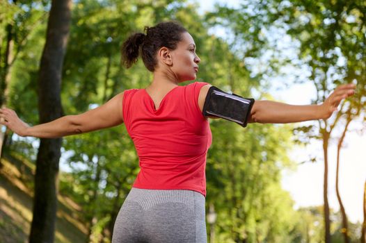 Confident determined beautiful fit woman, athlete in red t-shirt and gray leggings stretching out, enjoying a morning cardio workout and relaxation exercises in the forest park on a summer sunny day