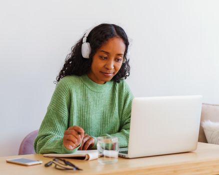 Young focused African-American female wireless studying on laptop in headphones while making notes in notebook. Concept of distance e-learning education at home. Taking online courses