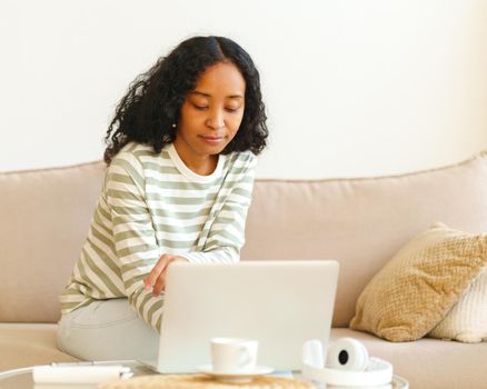 Young African-American female surfing the net on laptop while sitting on couch. Using digital device for remote work and e-learning. Concentrated and focused woman working at home