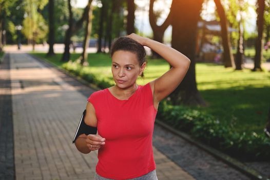 Determined middle-aged sportswoman in red t-shirt with smartphone holder tying ponytail while standing on path during morning jog along park. Cardio workout, active and healthy lifestyle concept