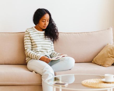 African-American woman in casual comfy clothes reading book while sitting on sofa in cozy living room. Home interior. Concept of maintaining emotional wellbeing. Having micro-moment and enjoying life