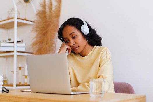 Bored and sleepy African-American female student in headphones attending online course on laptop at home. Fatigue and lazy millennial visiting distance class. Wireless watching lecture