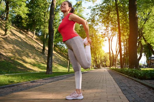 Side portrait of a middle-aged sporty woman stretching her leg muscles during a morning workout in a city park on a sunny warm summer day. Sports and a healthy lifestyle to keep fit