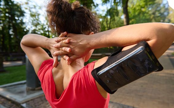 Close-up of a dark haired sportswoman with tanned skin wearing tight red t-shirt and a smartphone holder stretching out her body during morning workout in the city park on a warm sunny summer day