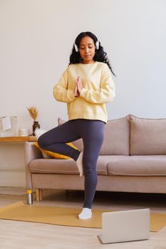 Young African-American female in sportswear and headphones standing in yoga tree pose at home while following fitness tutorial on laptop. Vertical. Maintaining physical and mental wellbeing