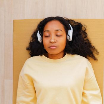 Tired African-American female lying down on mat with eyes closed and wireless listening to music while relaxing after training. Meditation practice. Concept of modern yoga exercises.