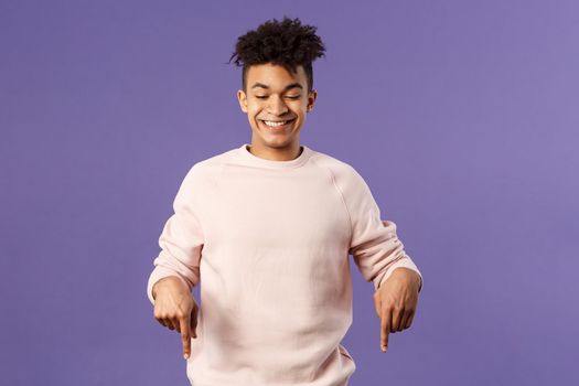 Portrait of cute enthusiastic young man found something really cool, pointing fingers down, happy smiling and peeking at bottom advertisement with pleased grin, purple background.