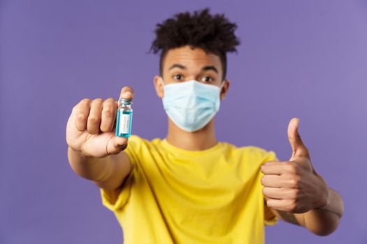 Close-up portrait of upbeat young hispanic man in medical mask holding ampoule with covid19 vaccine, coronavirus drug, show thumbs-up, getting shot at ambulance, standing purple background.