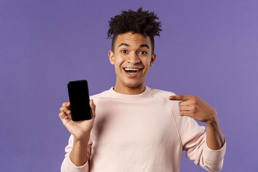 Portrait of excited, happy smiling man talking about new application or game feature, pointing at smartphone display, talking about mobile phone with amused cheerful expression.
