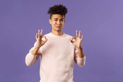 Portrait of skeptical young hispanic man rate something normal quality, show okay sign and grimacing judgemental, saying its not bad but average, standing purple background.