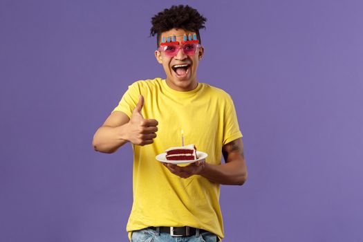 Celebration, party and holidays concept. Portrait of upbeat, cheerful young happy man celebrating birthday, wear funny sunglasses and hold b-day cake with lit candle, show thumb-up, recommend.