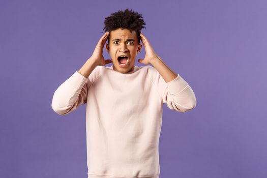 Portrait of scared and anxious young hispanic handsome man in panic, shaking hands nead head, screaming and gasping afraid, being in trouble, standing alarmed over purple background.