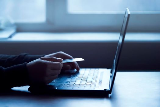 A man holds a bank card in his hand and types on a laptop keyboard, pays for purchases, makes online banking through an application, close-up view of his hands.