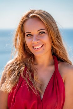 Smiling young woman in a red dress looks at the camera. A beautiful tanned girl enjoys her summer holidays at the sea. Portrait of a stylish carefree woman laughing at the ocean