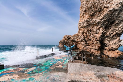 A woman in a blue jacket stands on a rock above a cliff above the sea and looks at the raging ocean. Girl traveler rests, thinks, dreams, enjoys nature. Peace and calm landscape, windy weather