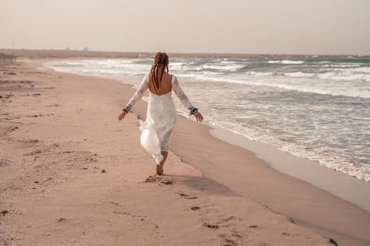 Model in boho style in a white long dress and silver jewelry on the beach. Her hair is braided, and there are many bracelets on her arms