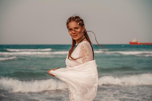 Model in boho style in a white long dress and silver jewelry on the beach. Her hair is braided, and there are many bracelets on her arms