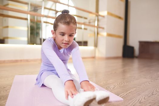 Adorable photo of little ballerina in a lilac dress and white tights stretching exercises in a hall with large mirrors.