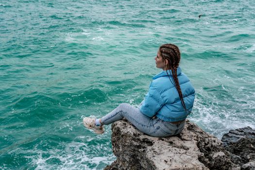 A woman in a blue jacket sits on a rock above a cliff above the sea, looking at the stormy ocean. Girl traveler rests, thinks, dreams, enjoys nature. Peace and calm landscape, windy weather