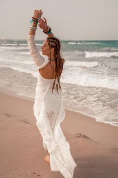 Model in boho style in a white long dress and silver jewelry on the beach. Her hair is braided, and there are many bracelets on her arms