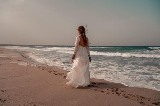 Model in boho style in a white long dress and silver jewelry on the beach. Her hair is braided, and there are many bracelets on her arms