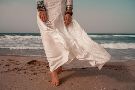 Model in boho style in a white long dress and silver jewelry on the beach. Her hair is braided, and there are many bracelets on her arms