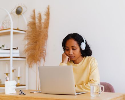 Sleepy and tired African-American woman in headphones studying online. Concept of e-learning at home on quarantine. Attending distance class. Home interior, pastel colors. Horizontal, copy space