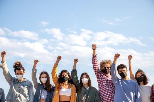 Group of multiracial protesters with fists raised up in the air wearing protective face mask. Activists protesting on the street. Copy space. Demonstration concept.