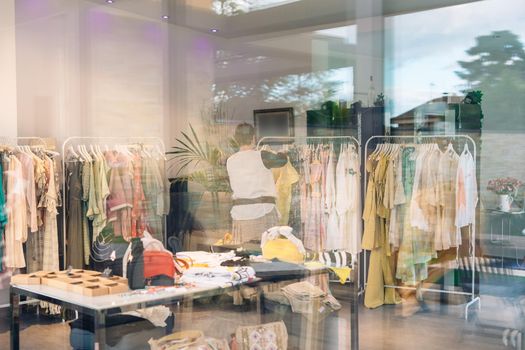 view from the outside of an elderly woman inside a clothes shop. shop window, shopping concept. woman selling clothes in her small shop. natural light from the window, sun rays, mirror, display with clothes, coat rack, window, clothes, Horizontal.